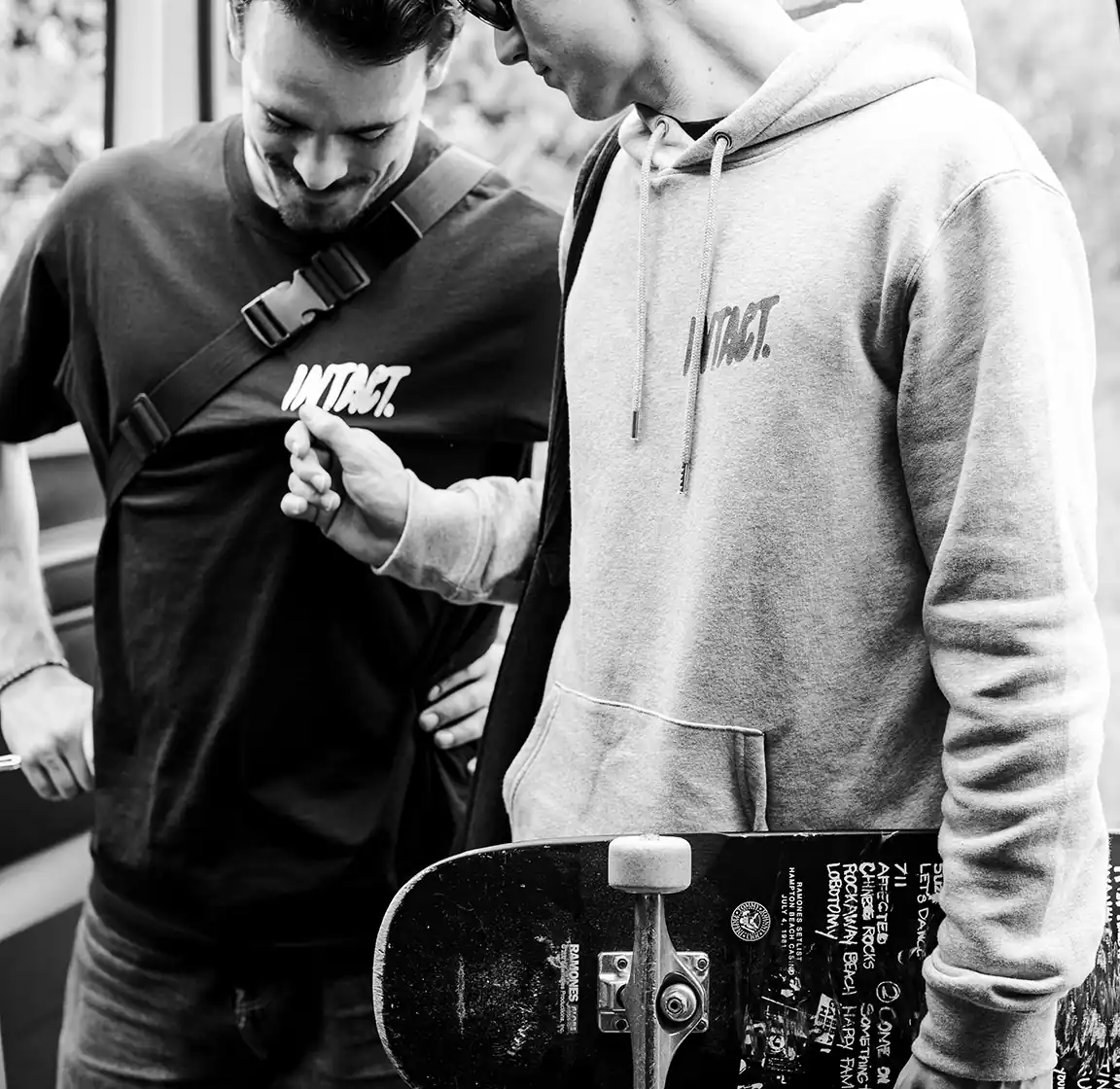 Deux jeunes hommes en tee-shirt et sweat de la marque Intact Something, l’un tenant un skateboard