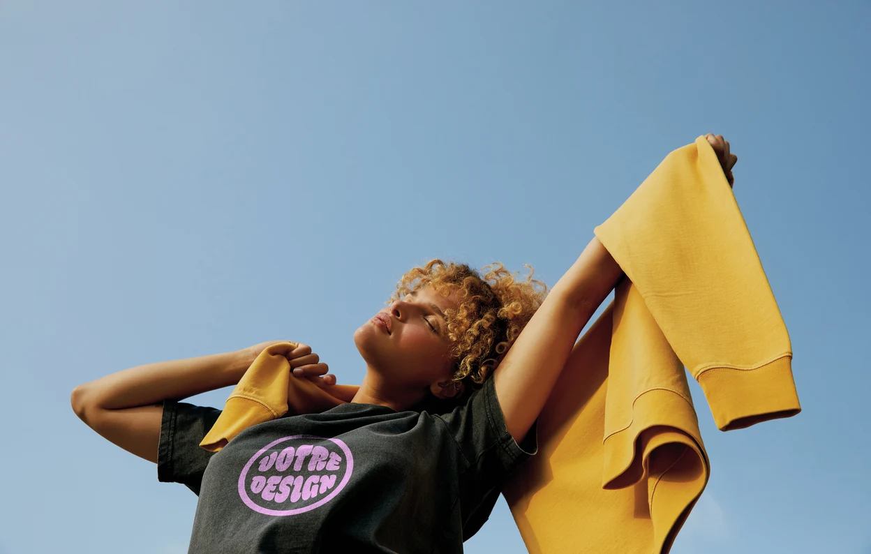 Femme portant un tee-shirt personnalisé, un sweat jaune sur les épaules, sous un ciel bleu